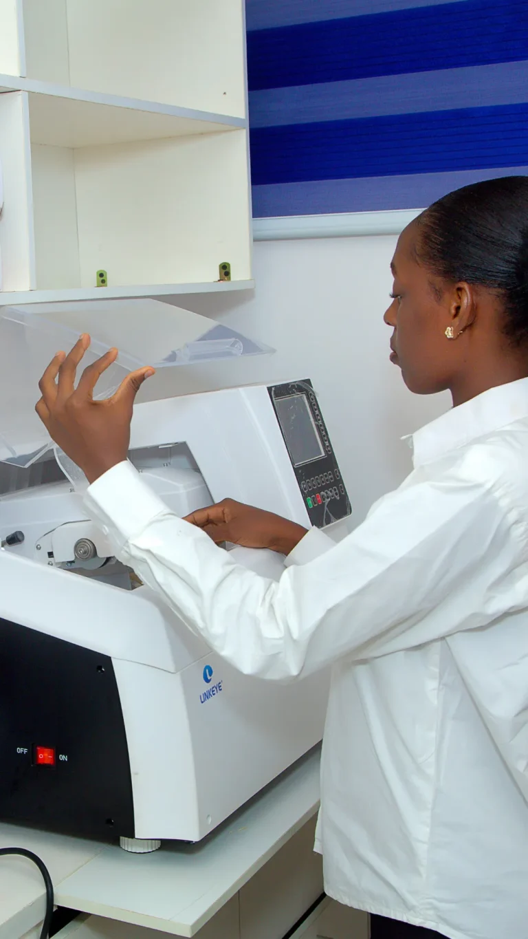 Optician using an automated lens edging machine (auto edger) to precisely cut and fit prescription lenses into eyeglass frames, providing fast on-the-spot glazing services at IrisCare Eye Clinic.